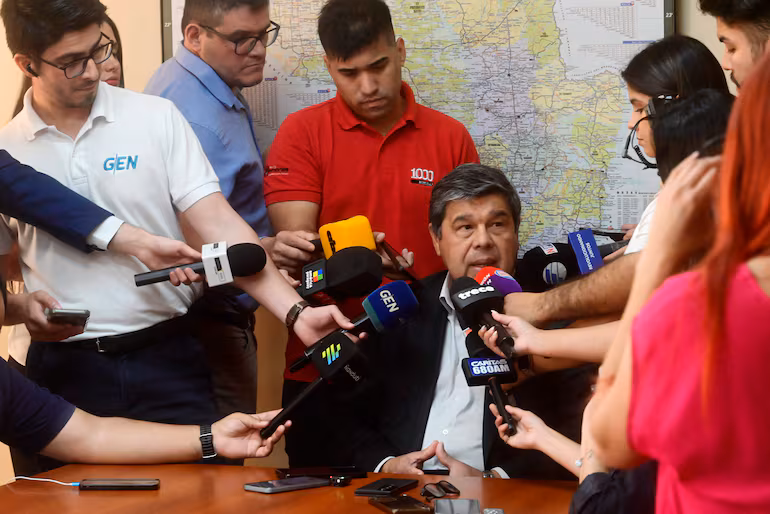 Hugo Arce en traje oscuro, sentado en mesa mientras responde preguntas de reporteros y cámaras en sala de prensa.